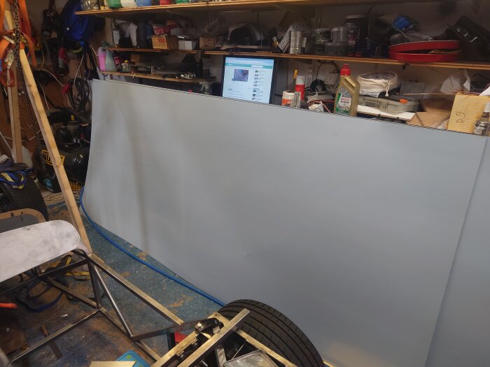 Verkstad med aluplåt lutad mot hylla, omgivande verktyg och byggmaterial, pågående fordonsprojekt, bildskärm i bakgrunden.