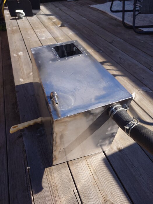 Svetsad metalltank med slanganslutning placerad på en träaltan i dagsljus.