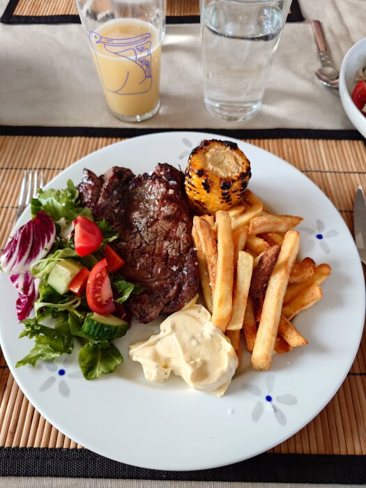 Tallrik med biff, pommes frites, bearnaisesås, grillad majs och sallad med tomat och gurka. Glas med dryck i bakgrunden.