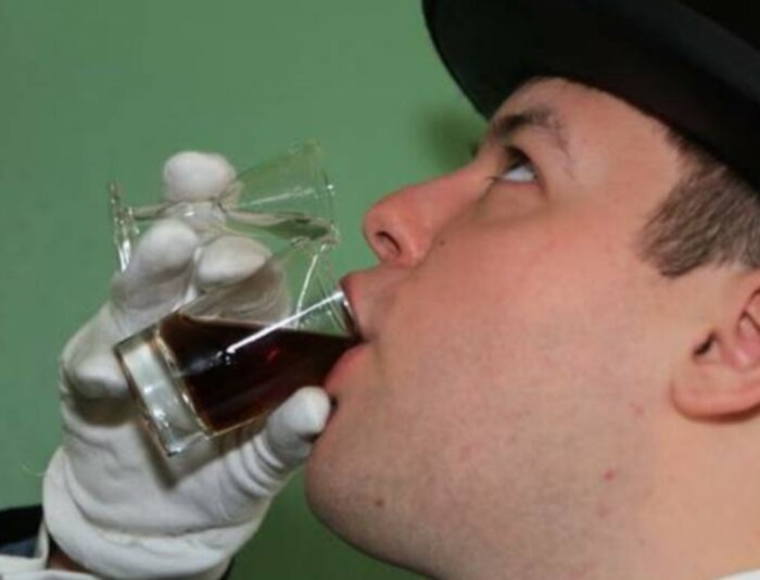 En person i hatt och vita handskar dricker en mörk vätska från ett shotglas mot en grön bakgrund.