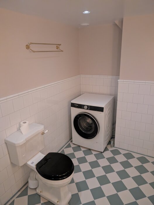 Modernt, nyrenoverat badrum med schackrutigt golv, vit tvättmaskin och toalett med svart sits. Ljusa väggar med vit kakelpanel och guldfärgad handdukshängare.