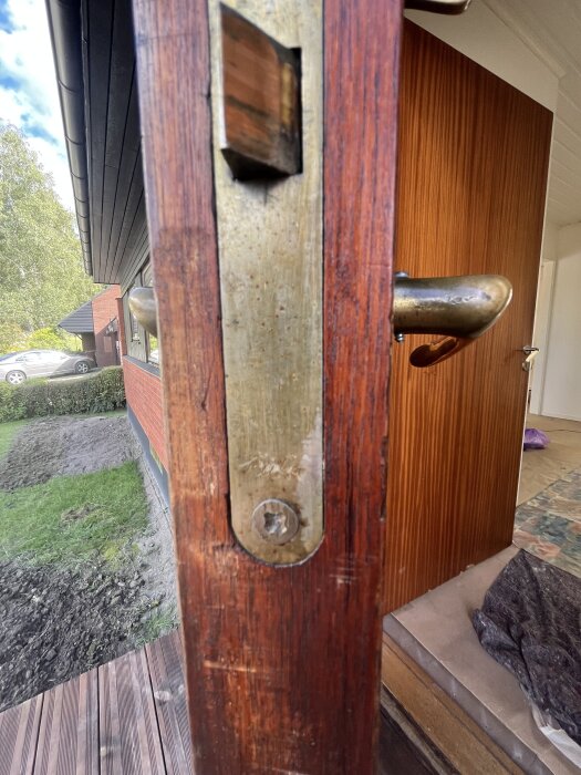 Närbild av ett brunt dörrlås med metallplatta på trädörr, sedd utifrån med grön trädgård och röd tegelbyggnad i bakgrunden.