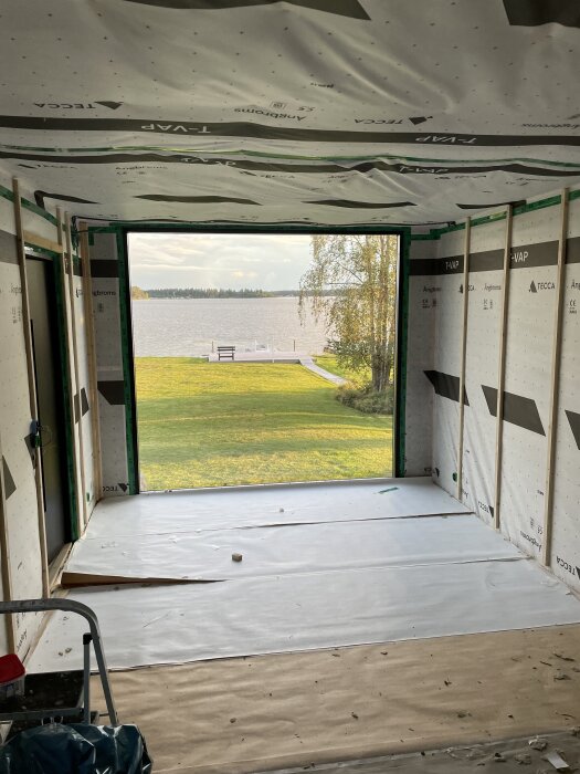 Byggarbetsplats inuti bastu med ångbroms och läkt på väggarna; utsikt mot en sjö och brygga genom en stor öppning.