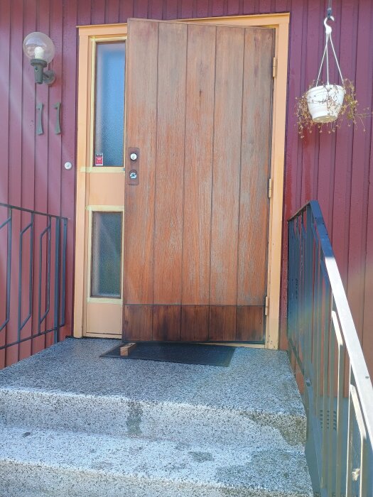 Dörr i trä efter teakrengöring och teakulja applicering, utan metalldetaljer. Vänsterhängd dörr med glasruta i orange karm, hängande kruka till höger.