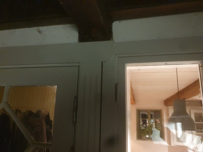 Kant av dörr med glasruta och synlig gångjärn, innerbelysning med ett hängande taklampa och en växt bredvid ett fönster i bakgrunden.