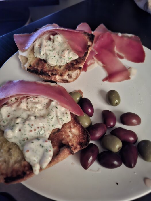 Ciabatta med Skagenröra och skiva lufttorkad skinka på en tallrik, omgiven av gröna och svarta oliver.
