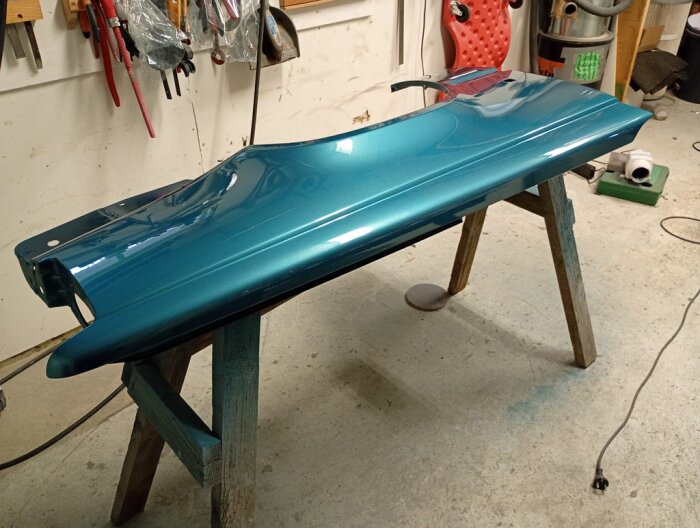 Blå bilpanel, nyligen klarlackad, placerad på träbockar i ett garage med verktyg på väggarna.