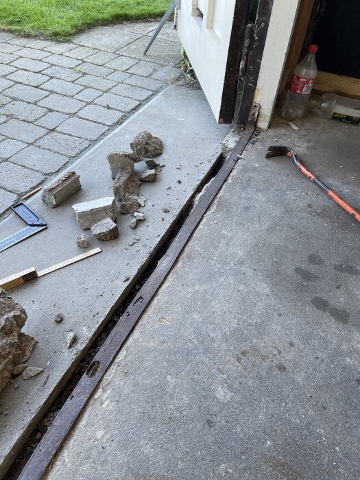 Garageinfart med sprickor och rostig L-profil, bitar av trasig cement och verktyg i förgrunden, gräsmatta i bakgrunden.