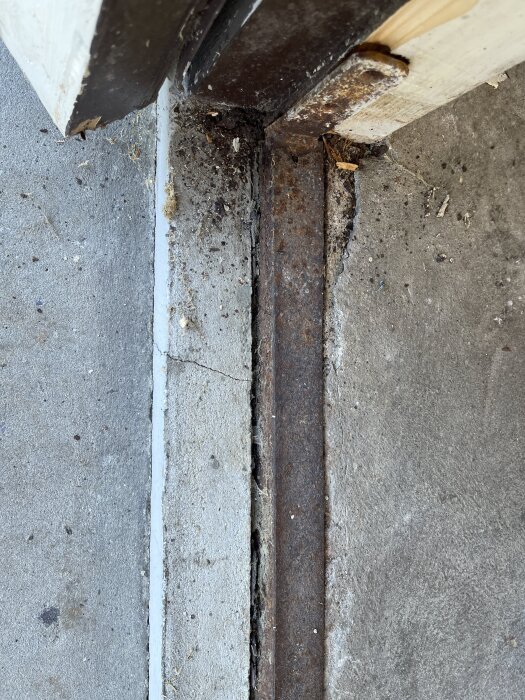 Rostig och sprucken garageinfart med synlig L-profil vid kanten, omgiven av betong och skadad trästruktur.