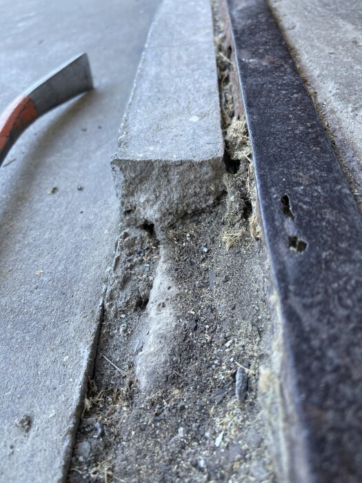 Sprucken garageinfart med sliten L-profil i metall och trasig betong, synligt slitage och rost vid kanterna, verktyg synligt i bakgrunden.