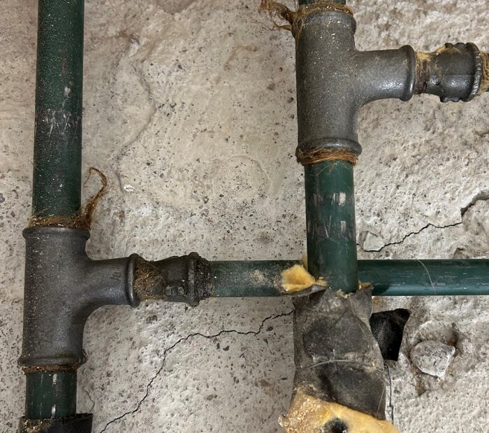 T-rör i metall med gröna rör och tätningar. Rören är del av ett äldre radiatorsystem, med smalare rör som sticker ut åt höger.