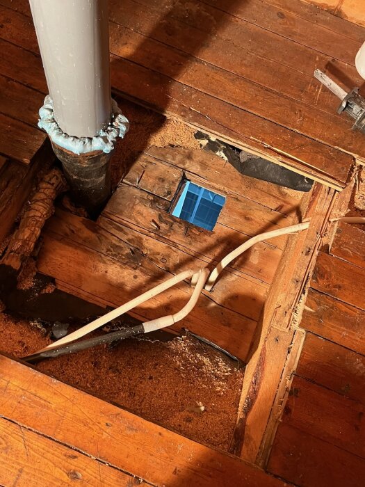 Isärsågad eternitkanal i ett trägolv, omgivet av sågspån och mindre rör. En tigersåg ligger till höger på bilden.