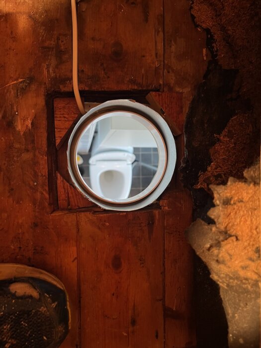 Genomskärning av trävägg med ventilationskanal där man ser in i ett badrum, del av sko och isoleringsmaterial synligt.