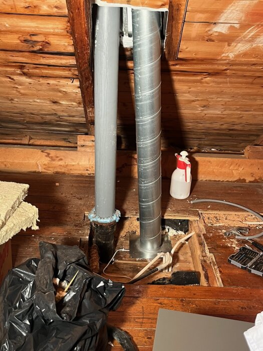 Två ventilationsrör monterade på en vind, ett grått spirorör och ett metalliskt, med isolering och verktyg runtomkring.
