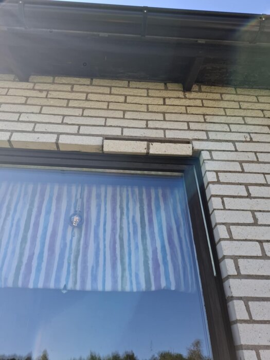 Tegelvägg med två lösa tegelstenar ovanför fönster, stenen hänger delvis ur murverket.