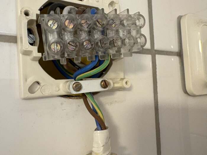 Öppnad kopplingsdosa med färgade kablar, inklusive brun fas, synliga mot vit kakelvägg i samband med tvättmaskinsinstallation.