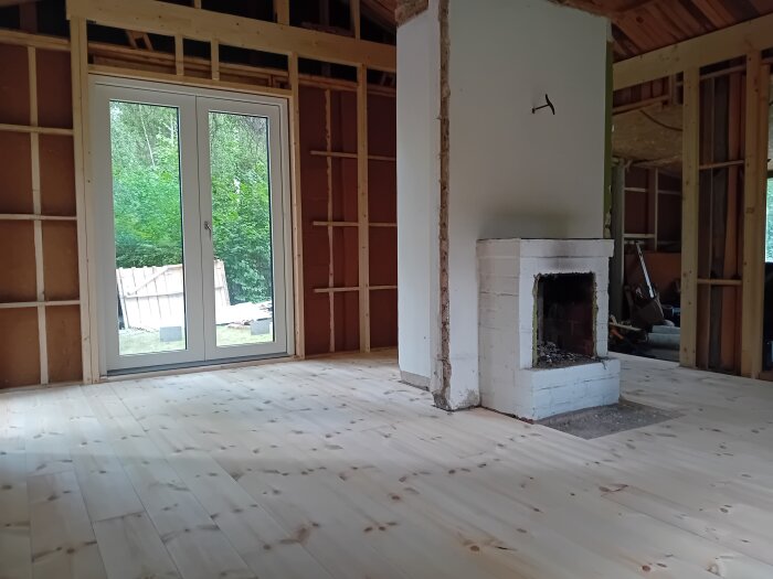 Ljust furugolv i ett rum med oanalyserade väggar och en vit öppen spis, stora fönsterdörrar med utsikt mot trädgård.