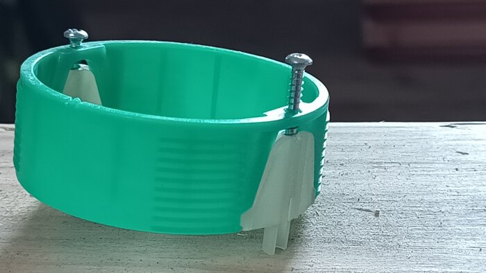 En grön plastkomponent med två skruvar monterade i det övre skiktet, placerad på en träyta.