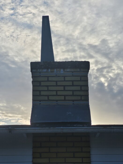 Skorsten i tegel med en löst sittande metallpipa ovanpå, mot en mulen himmel i bakgrunden.