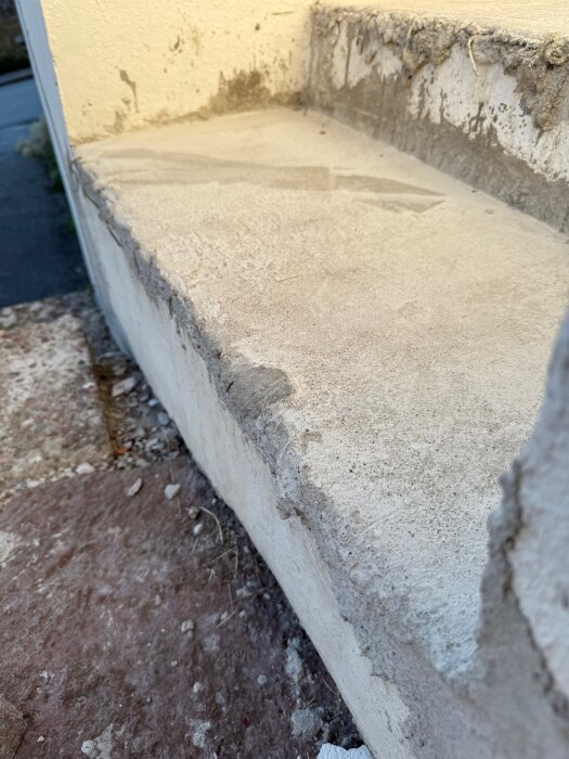 Gjutet cementsteg i trappa under konstruktion med ojämnt lager av cement och armering synlig, nära intill vägg med sliten yta.