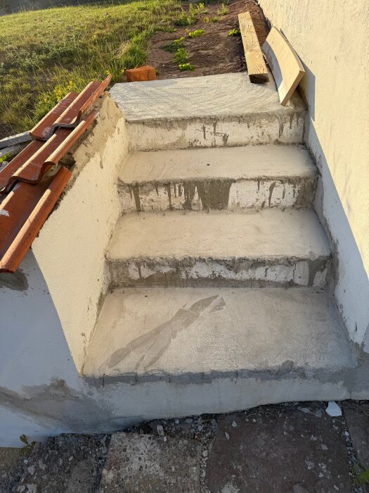 Ofärdig cementtrappa utomhus med ojämn yta och några lösa tegel- och träbitar vid sidan. Gräs och jord syns runt om.