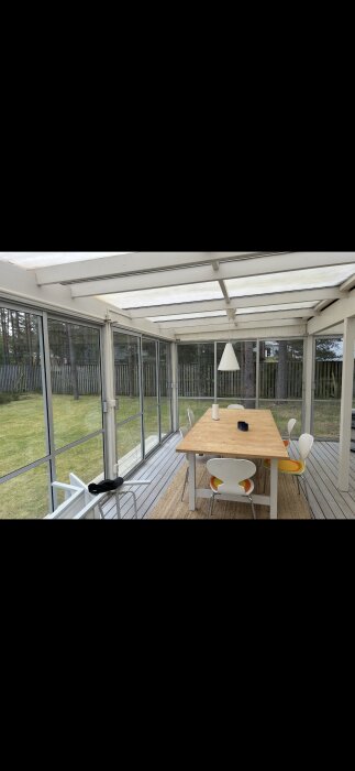 Ett oisolerat uterum med kanalplasttak, glasväggar och bord med stolar. Utsikt över en gräsmatta och skog.