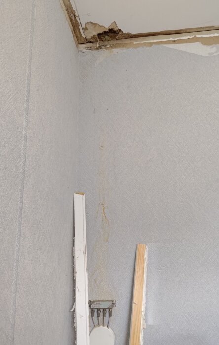 Fuktskador i ett hörn av ett rum, med skadat tak och väggar, samt synliga rör och bitar av lös inredning.