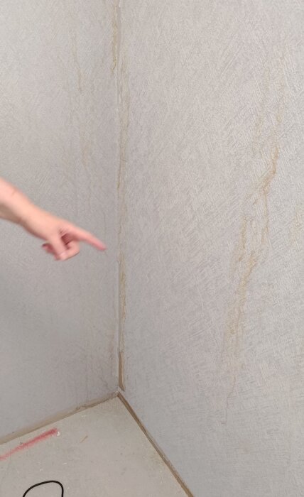Finger pekar på vattenskador i skafferihörn, med märken längs väggen och en ledning synlig på golvet.