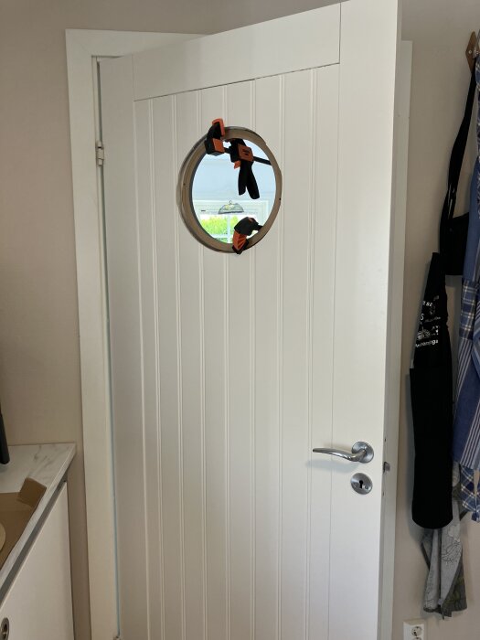 Omlackad vit dörr med runt fönster, säkrat med klämmor.