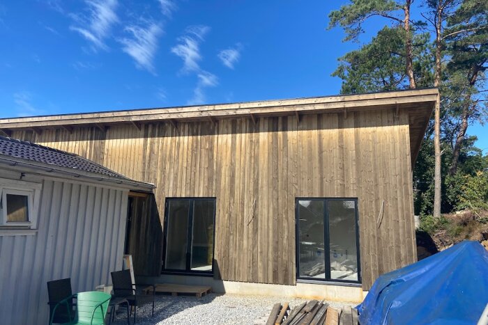 Träfasad på hus med skiftande färgtoner och skugga från överhäng. Några små mörka fläckar, omgivningar med träd och klar himmel.