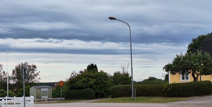 Landsväg i en förort med gatlykta, buskar och ett gult hus under en molnig himmel. Trafikskylt visar hastighetsbegränsning 30.