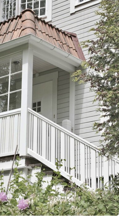 Vitt träräcke med spjälor på verandatrappa, spjälorna placerade vid sidan av räcket istället för mitten, gröna växter i förgrunden.