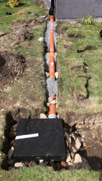 Orange markrör installerat i schakt med dränering, omgivet av grus och stenar. Synlig förbindelse med stuprör.