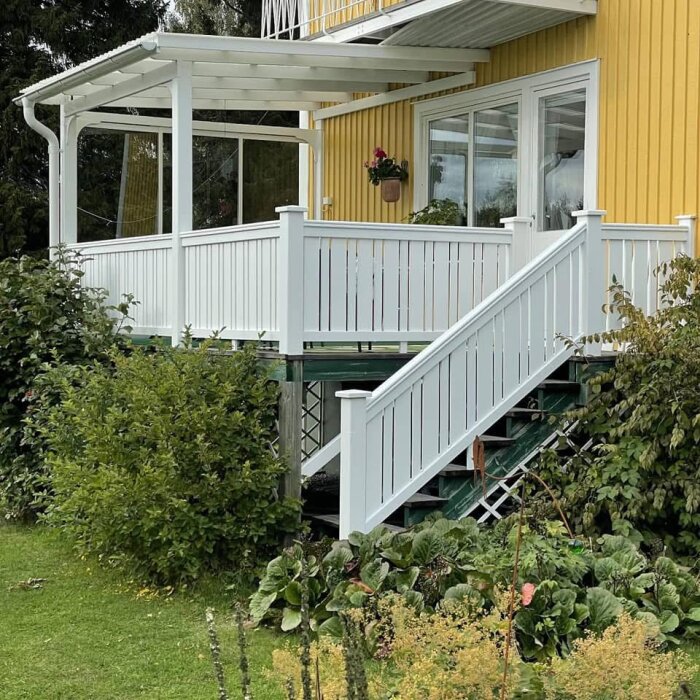 Vitmålat räcke med trappa intill ett gult hus, del av en terrass med grönska runtom.