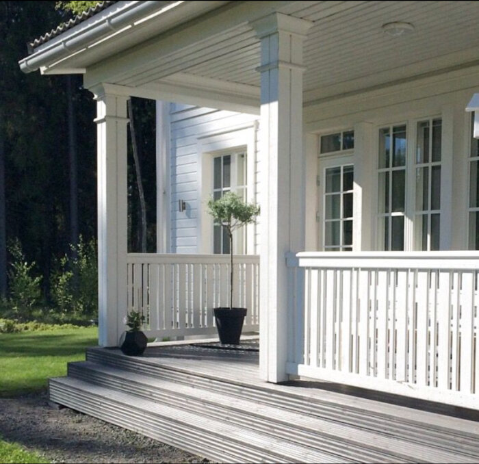 Vitt verandaräcke med pelare och krukväxter på en träterrass framför ett vitt hus.