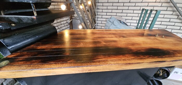 Utomhusbarbord av trä, bränt och behandlat med hantverksolja, står på en veranda med belysning och tegelvägg i bakgrunden.