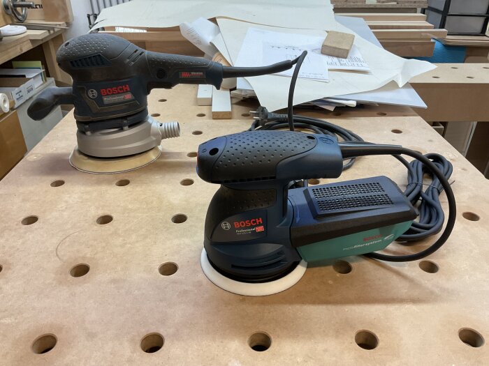 Två Bosch slipmaskiner, GEX 125-150 AVE och GEX 125-1 AE, står på ett arbetsbord i en verkstadsmiljö med papper och verktyg.