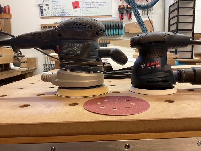 Två Bosch slipmaskiner på en arbetsbänk i en verkstad, en GEX 125-150 AVE och en GEX 125-1 AE, med en sliprondell emellan.