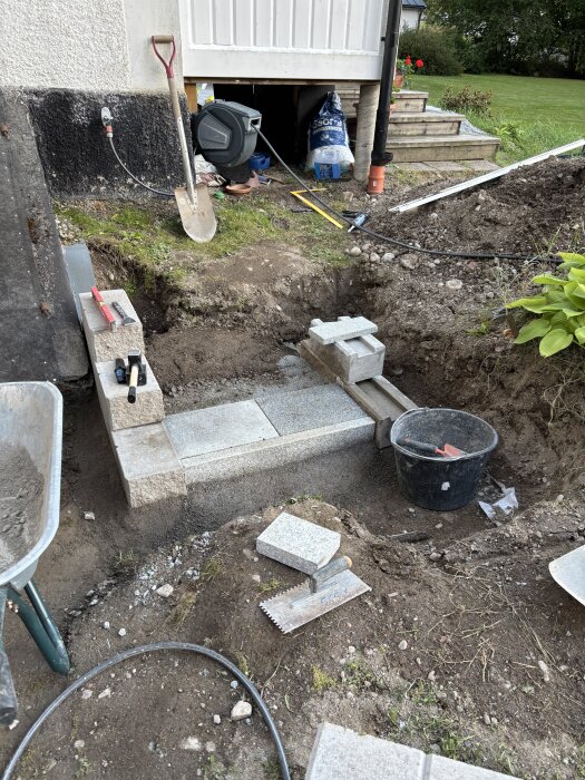 Byggprojekt av stentrappa med verktyg som murslev och hink, förberett för uteköksdel på veranda. Schaktad jord och byggmaterial syns.