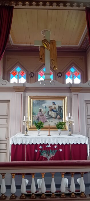 Kyrkligt altare med kors, målning av två personer vid en brunn och ljusstakar. Bakom syns färgglada glasfönster med röd och blå dekoration.