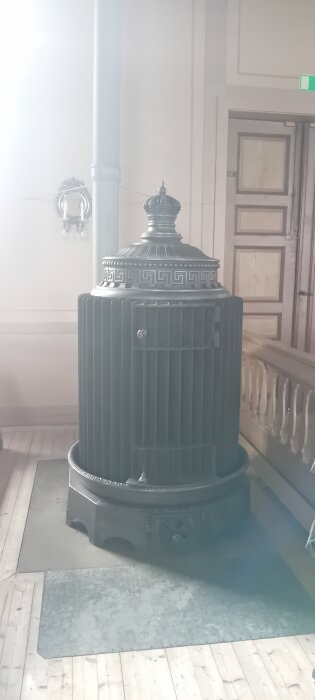 Gammal kamin med dekorativt mönster och krona på toppen, står på trägolv bredvid en vit vägg med dörr och väggsconce.