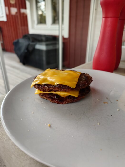 Två staplade hamburgarbitar med smält ost ovanpå en vit tallrik, i bakgrunden syns en röd pepparkvarn och ett rött hus.