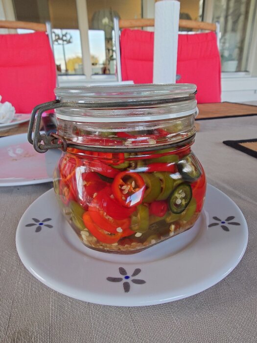 En glasburk med picklad chili i olika färger, placerad på en dekorerad tallrik på ett bord med röda stolar i bakgrunden.