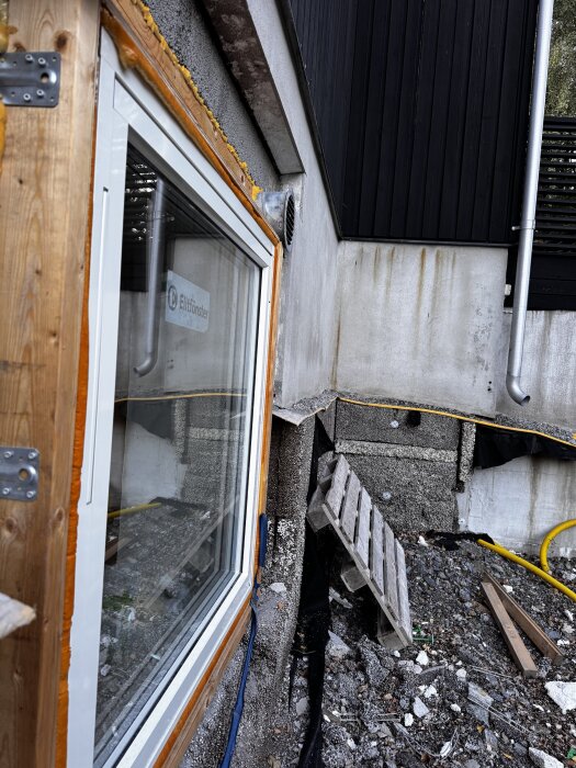 Sutteränghus med nytt fönster monterat, synlig ventil på fasad, lecablock utan puts, träskivor, och byggmaterial på marken.