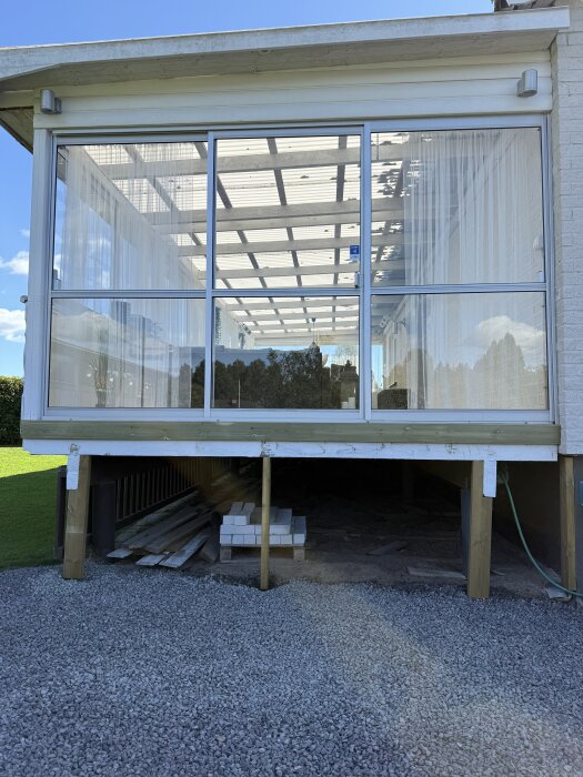 Uterum med glasväggar och tak, upphöjt på pelare, omgiven av grus och trämaterial under som förbereds för altanprojekt.