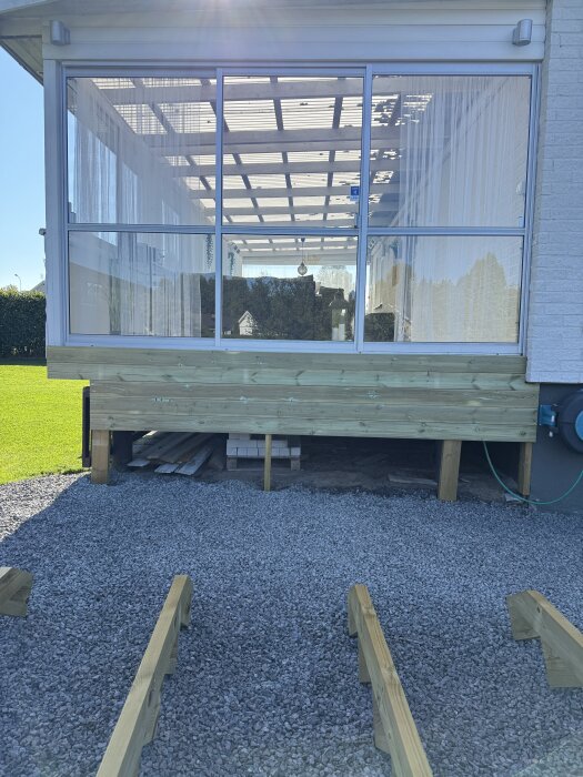 Byggarbete vid altan, där nedre delen kläs in med liggande trall. Grus och träreglar på marken förbereder platsen för ny trappa.