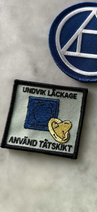 Textilmärke med budskapet "Undvik läckage, använd tätskikt" i svart text, med blå fyrkant och gul illustration.