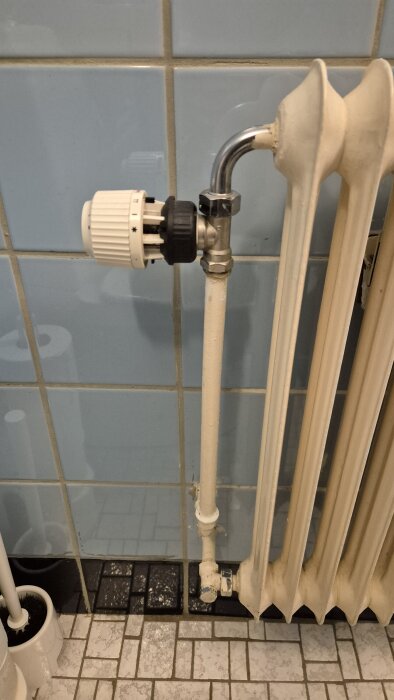 Element med termostat och rörkopplingar i ett badrum med blå kakelvägg och klinkergolv.