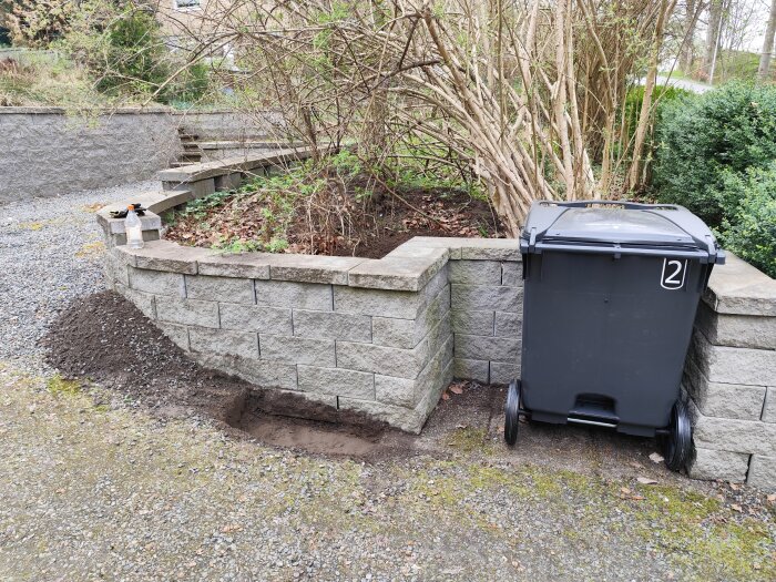 Grå soptunna vid nybyggd stenmur och grusväg, omgiven av buskar och träd. Spade och sprayflaska på muren.