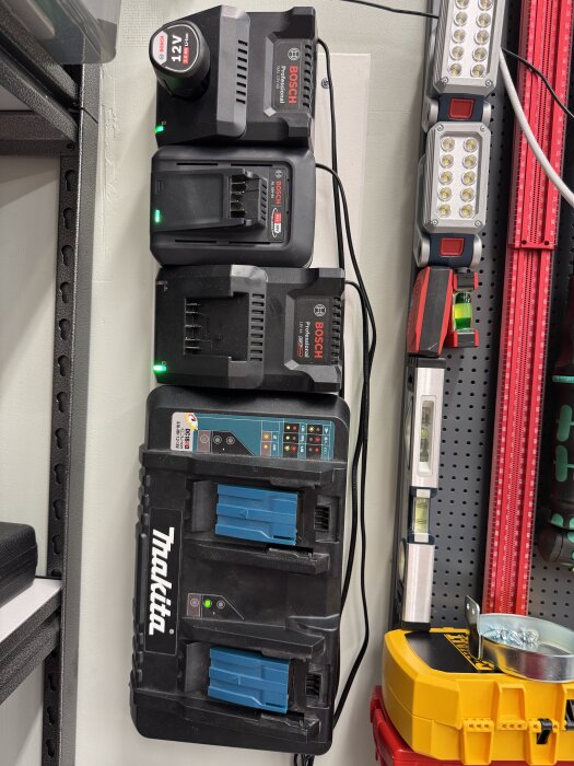 Batteriladdare monterade på väggen: 12v, två 18v Bosch (en grön och en blå), och en 18v Makita, omgivna av verktyg och utrustning.